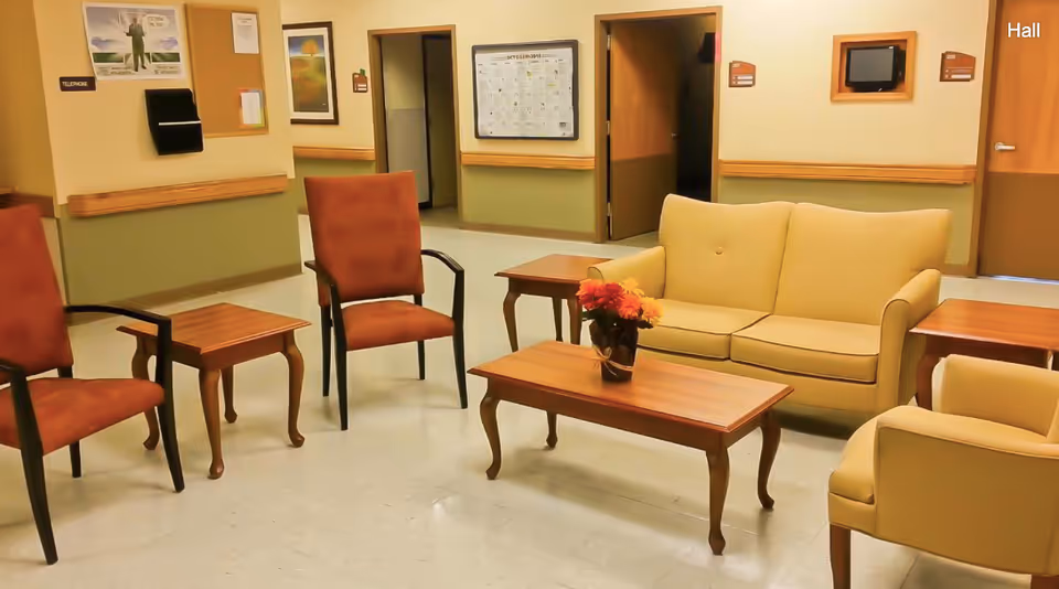 A seating area in a hallway of Deanwood Rehabilitation And Wellness Center featuring a beige loveseat, beige armchair, two red cushioned chairs, and wooden coffee and side tables. A small vase with flowers is on the coffee table. The walls have framed pictures, a calendar, and room number signs.
