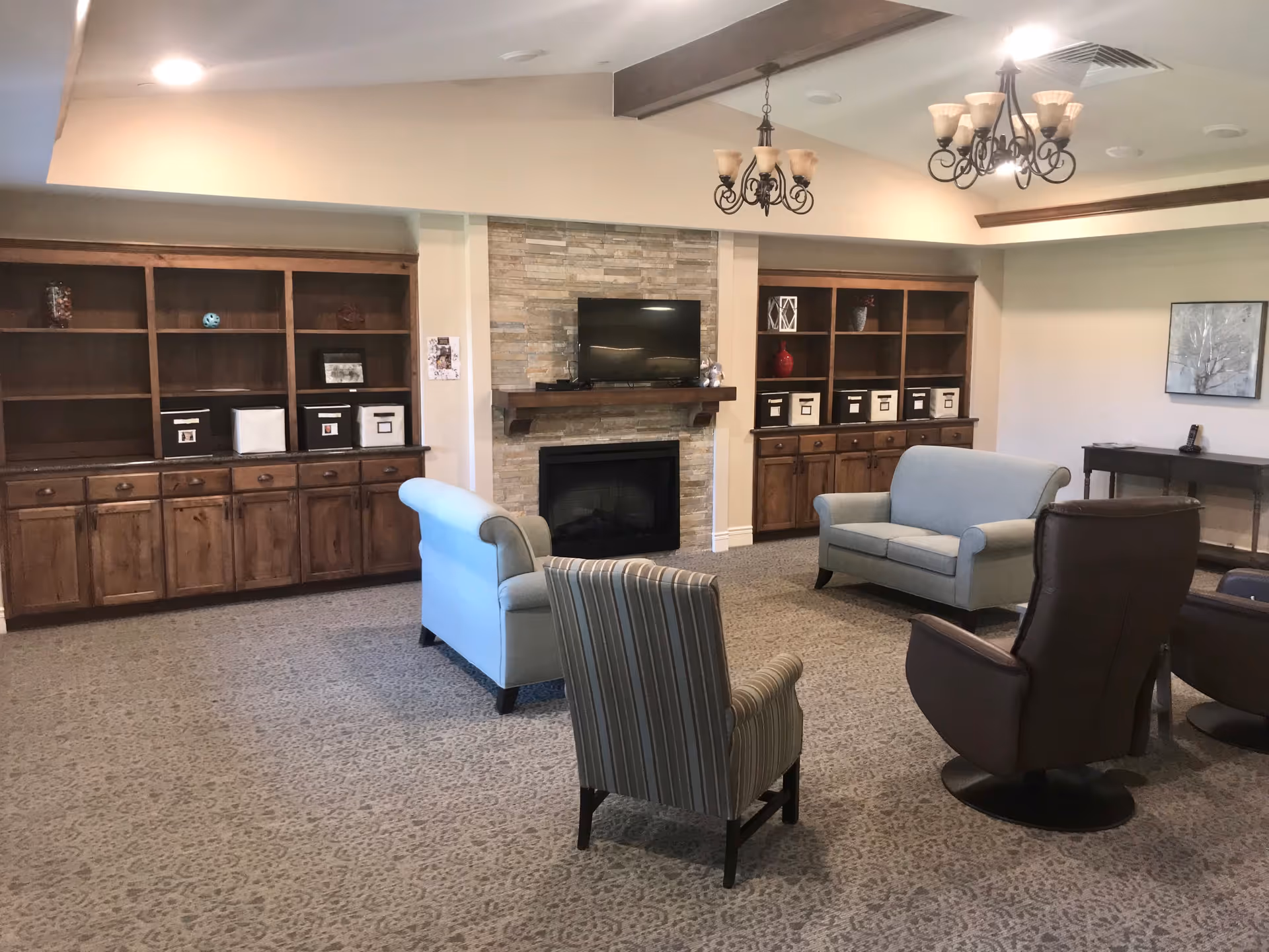 A cozy common lounge with sofas and armchairs arranged around a stone fireplace and TV, flanked by wooden built-in shelving and chandeliers.