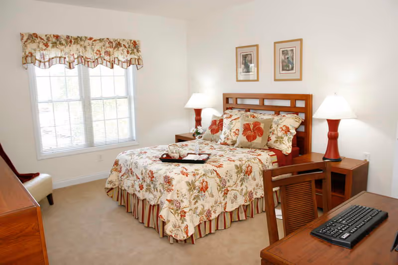 Bright furnished bedroom with a floral bedspread, wooden headboard and furniture, bedside lamps, and a window with blinds.