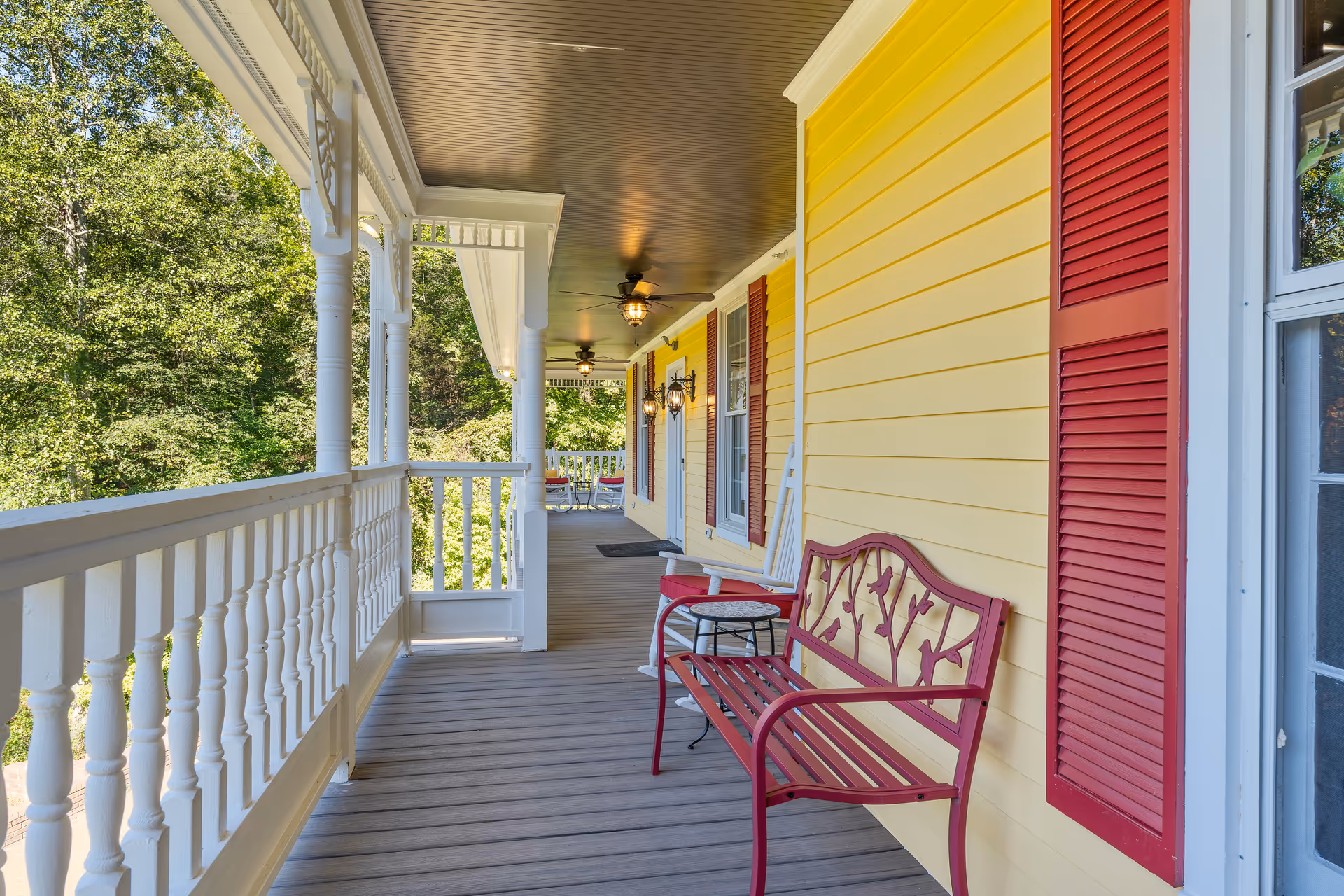 A yellow porch of a senior living facility with red shutters, white railing, and seating.