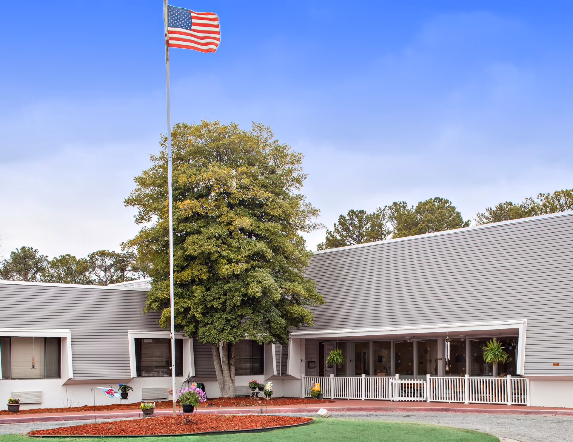 Exterior view of a single-story nursing and healing center building with gray siding and white trim. There is a large tree in front of the building, an American flag on a tall flagpole, and a small garden area with mulch and potted plants. The entrance has a white railing and hanging plants.