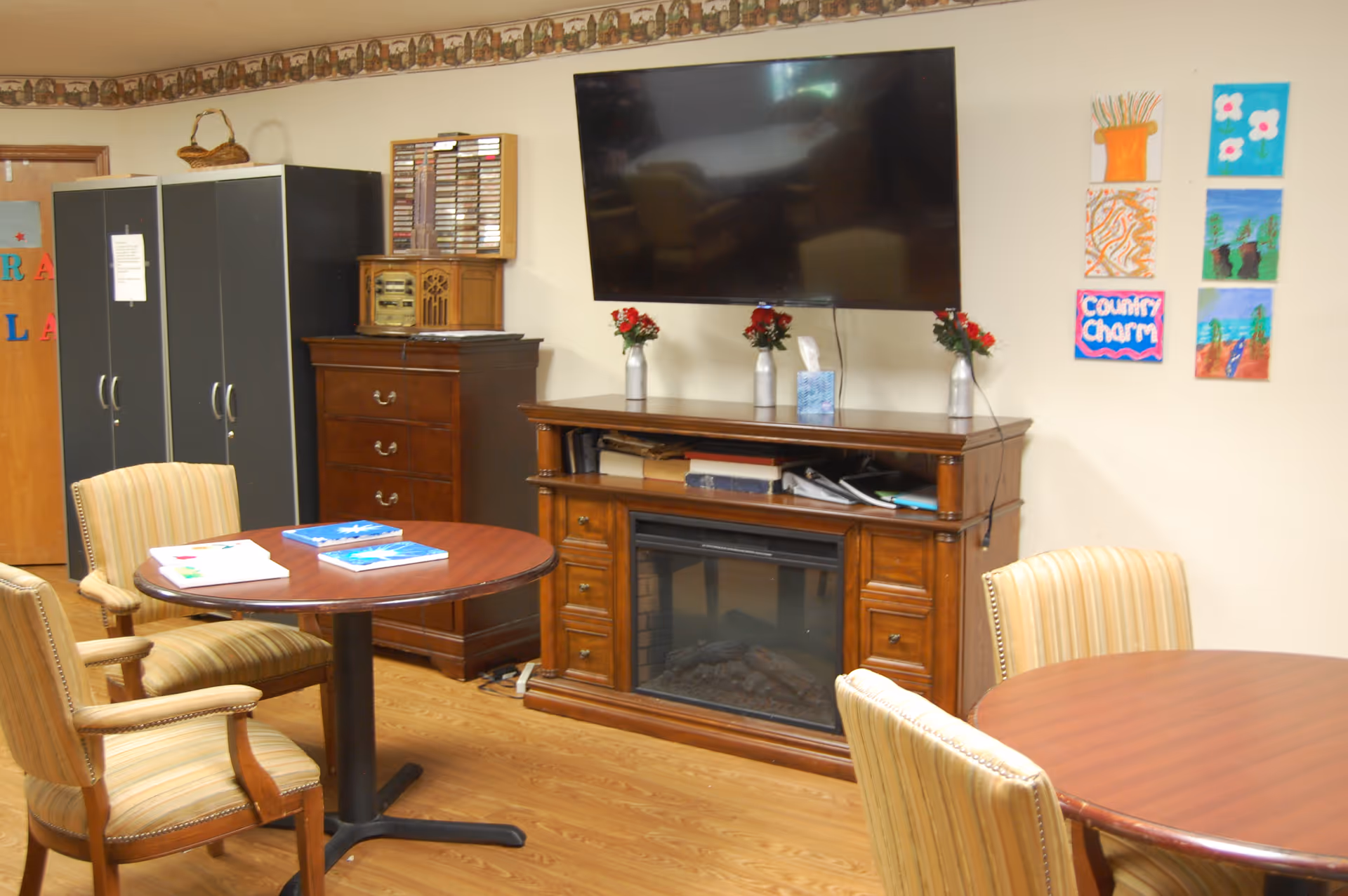 A cozy common area with two round wooden tables surrounded by striped upholstered chairs. A wooden cabinet with a built-in electric fireplace is topped with three small vases of red flowers and a tissue box, and a large flat-screen TV is mounted on the wall above it. On the wall to the right, there are six colorful paintings, one of which says 'Country Charm'. In the background, there is a tall black cabinet and a wooden chest of drawers with a vintage radio and cassette tapes on top. The room has wood flooring and a decorative border near the ceiling.