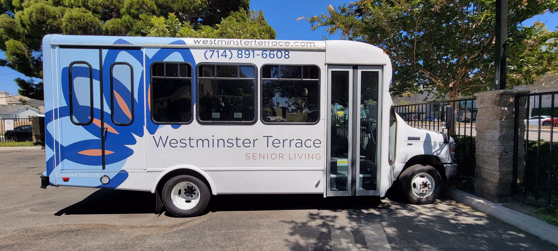A white shuttle bus parked outdoors with large blue and orange floral graphics on the side. The bus has the text 'Westminster Terrace Senior Living' along with the website 'westminsterterrace.com' and phone number '(714) 891-6608' displayed on its side. The bus is parked next to a fence with trees and a clear blue sky in the background.