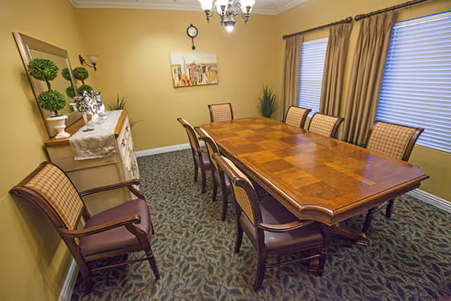 A dining room with a large wooden table surrounded by eight cushioned chairs. The room has beige walls, a patterned carpet, two windows with closed blinds and brown curtains, a chandelier hanging from the ceiling, a wall clock, a painting, and a sideboard decorated with plants and silver ornaments.