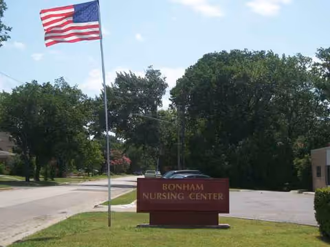 American flag beside a Bonham Nursing Center sign on a grassy, tree-lined exterior.