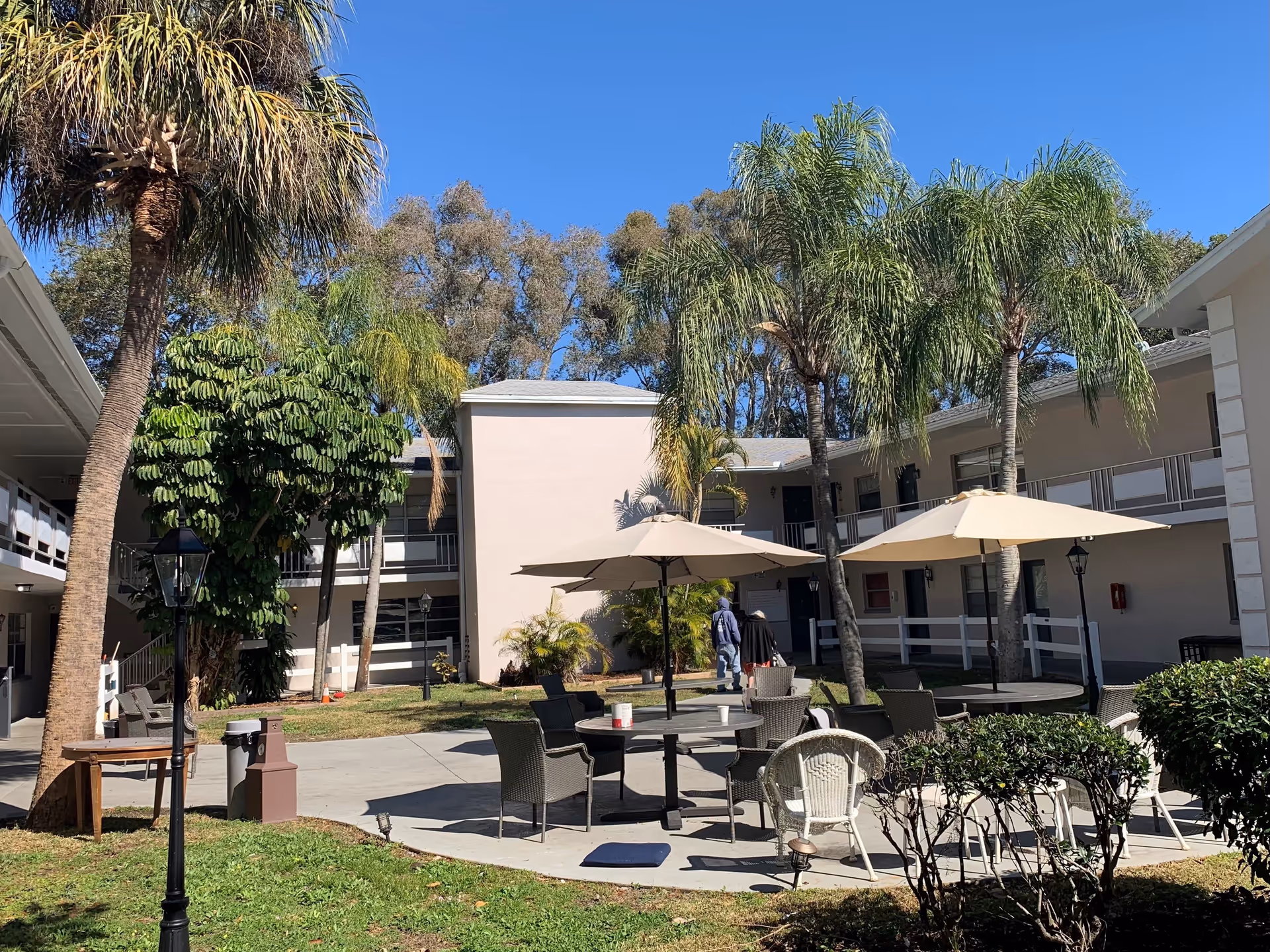Outdoor courtyard area at Midway Manor Assisted Living Facility with palm trees, patio tables with umbrellas, chairs, and two people walking near the building under a clear blue sky.