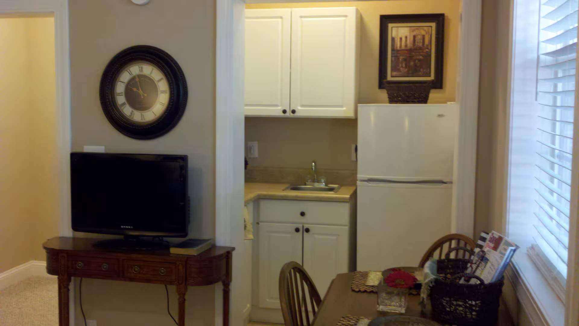 Interior view of a small kitchen area with white cabinets, a sink, and a white refrigerator. To the left, there is a wooden table with a flat-screen TV on it and a large round wall clock above. On the right side, there is a dining table with chairs, a basket with magazines, and a window with blinds.