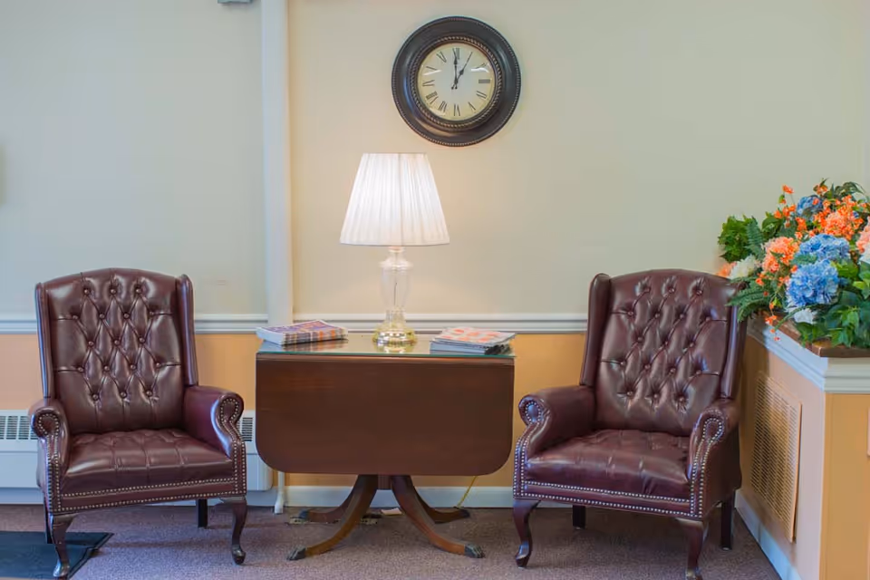 Two tufted brown leather armchairs flank a wooden side table topped by a lamp beneath a wall clock, with a floral arrangement on the right.