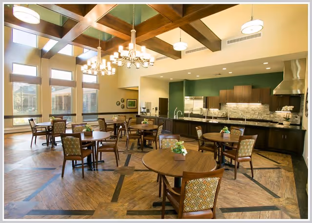 Spacious dining area with round wooden tables and chairs, chandeliers, and an open kitchen and service counter in the background.