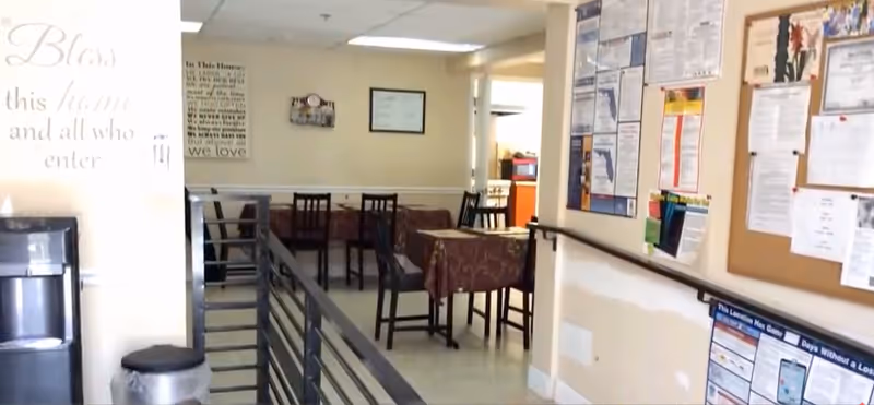 Interior view of a dining area in a retirement home with several tables covered with patterned tablecloths and chairs. On the right wall, there is a bulletin board filled with various notices and papers. On the left side, there is a water dispenser and a trash can. The walls are light-colored, and there are framed signs and decorations visible.
