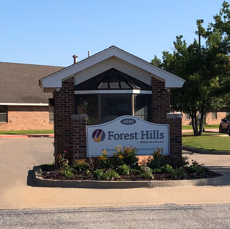 Entrance sign for Forest Hills Assisted Living, a senior living community by MGM Healthcare, set in front of a small brick structure with a landscaped flower bed and trees in the background.