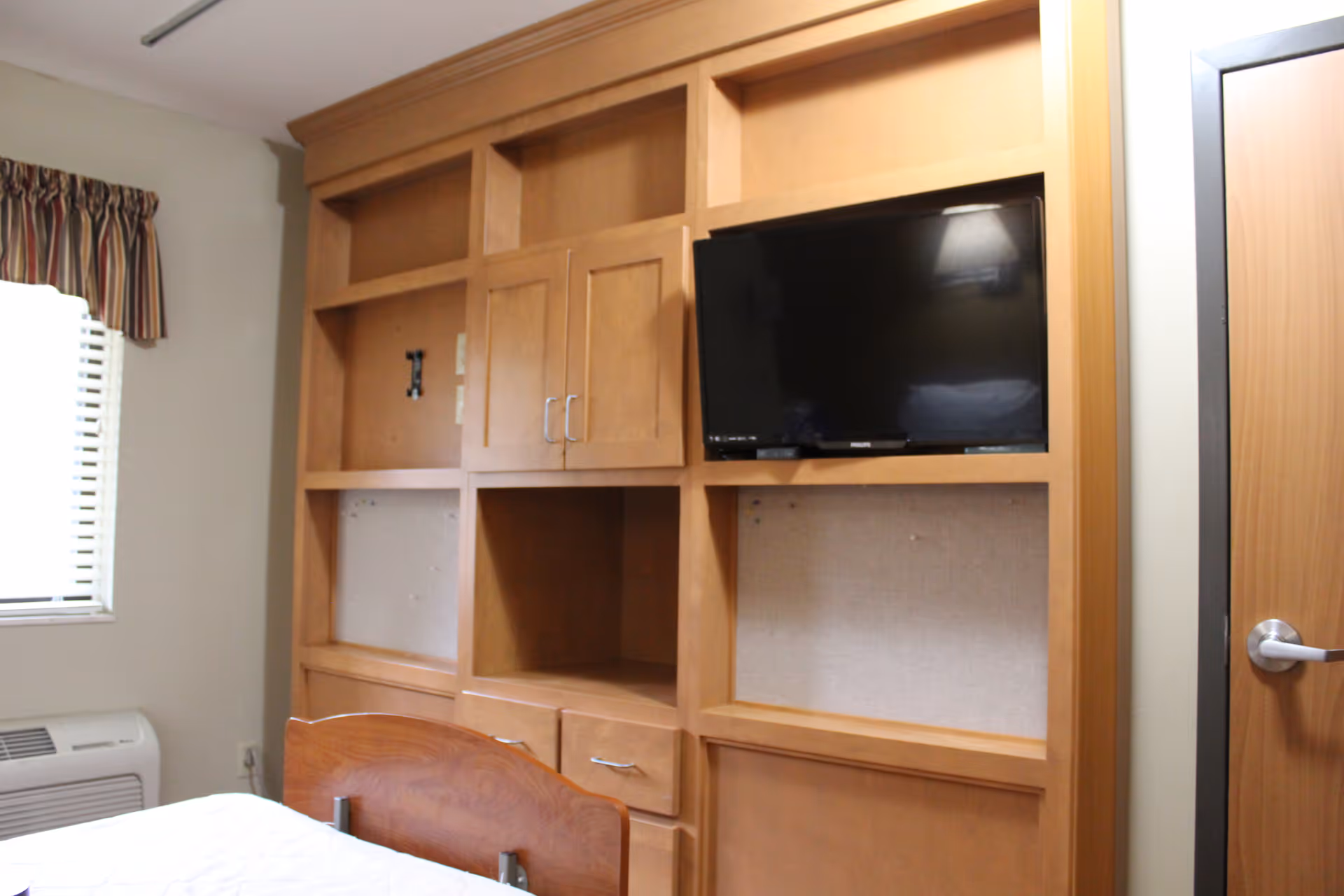 Interior view of a bedroom featuring a wooden built-in shelving unit with cabinets and open shelves, a mounted flat-screen TV, a window with striped curtains, a bed with a wooden headboard, and a door with a silver handle.