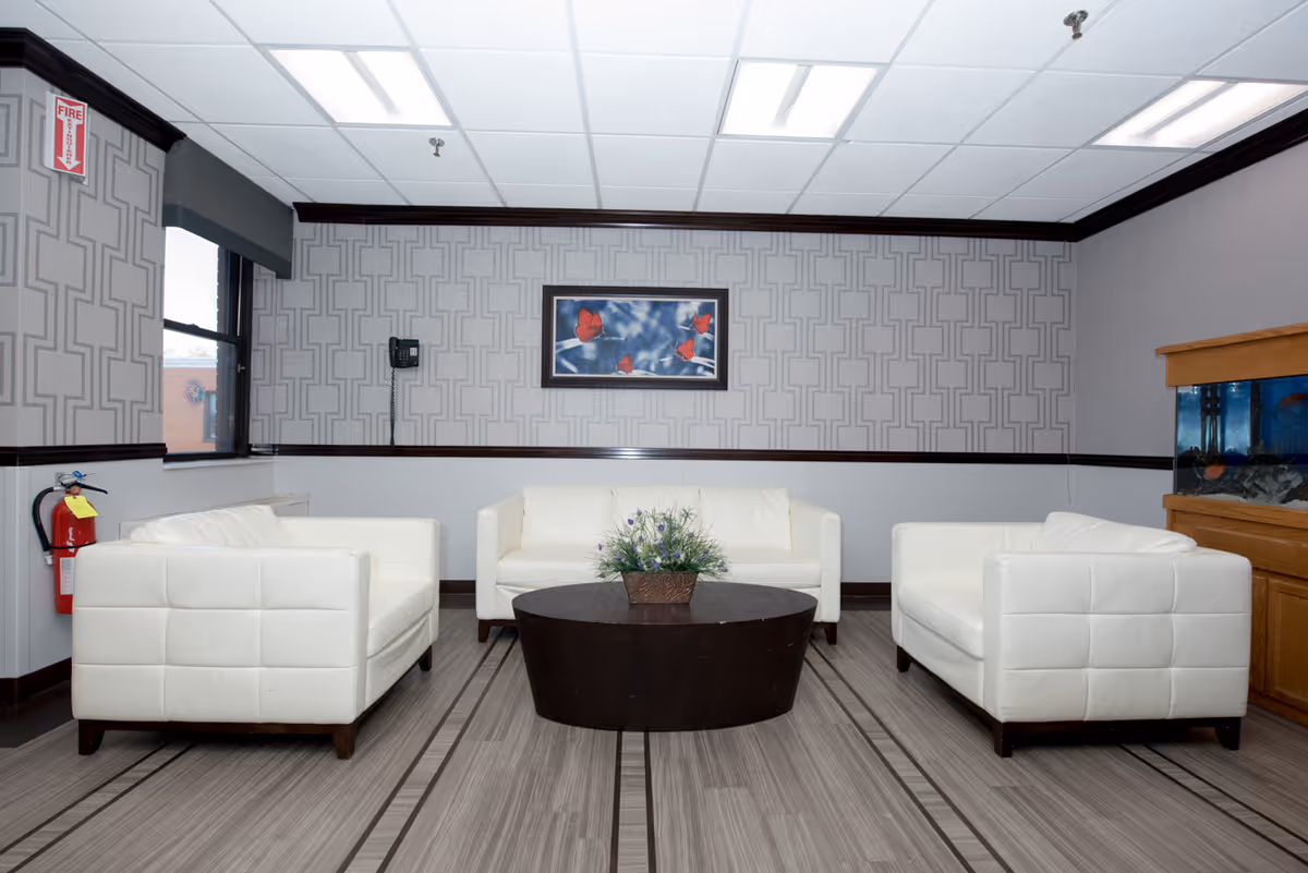 A seating area in a room with three white cushioned armchairs arranged around a dark round coffee table with a floral centerpiece. The walls have a geometric patterned wallpaper with a framed picture of red butterflies on a blue sky background. There is a fire extinguisher mounted on the left wall and a fish tank on a wooden cabinet on the right side of the room.