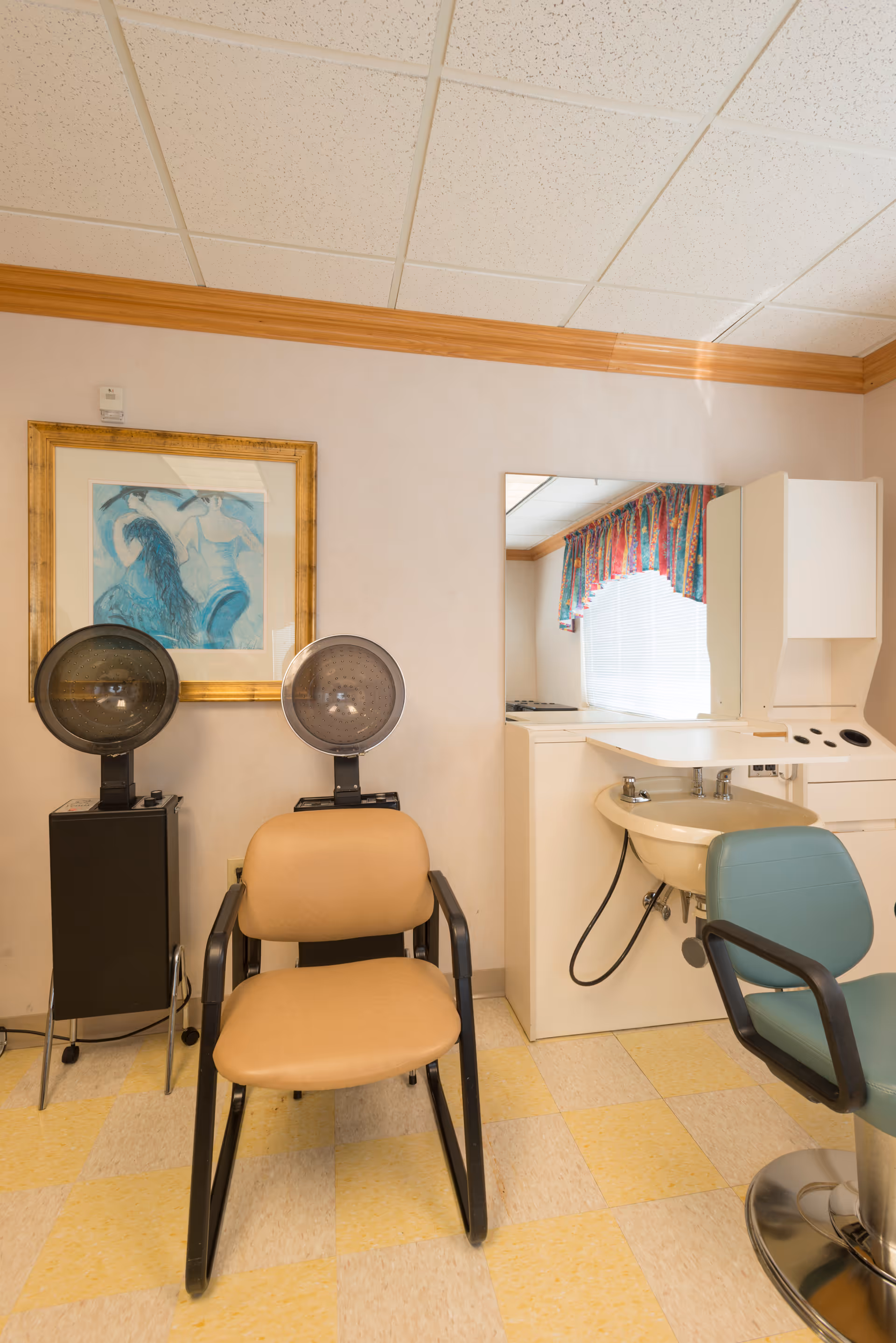 Interior of a hair salon area in a senior living facility with two salon chairs, two hair dryers on stands, a sink with a mirror above it, and a colorful valance on the window.