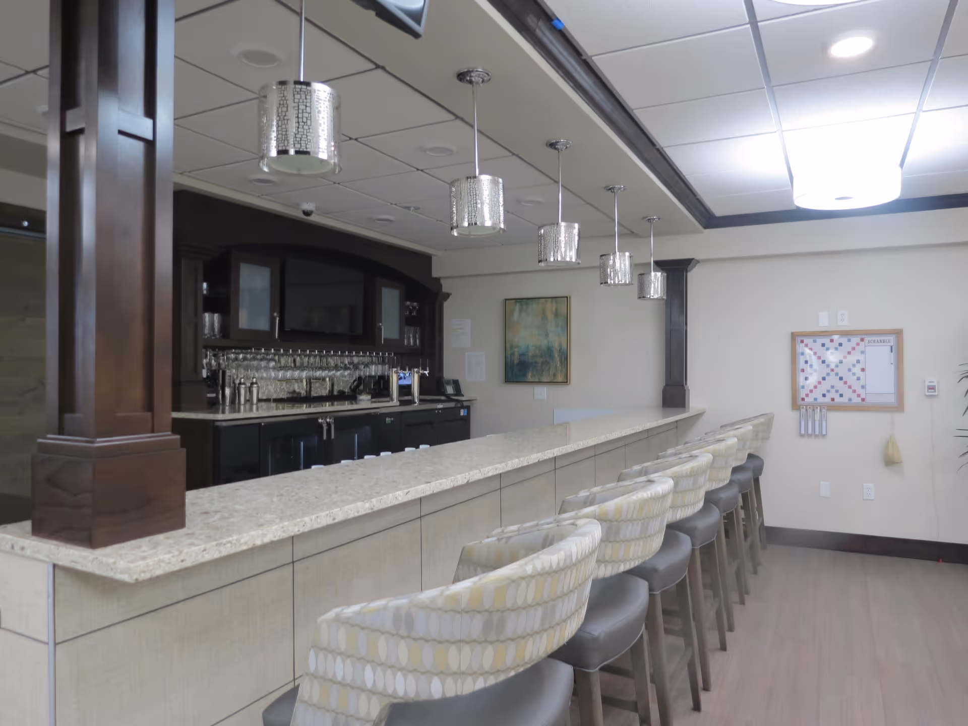 Interior view of a bar area with a long countertop and several cushioned bar stools lined up in front. The bar has dark wood cabinetry with glass doors, hanging pendant lights above, and a wall-mounted Scrabble board on the far wall.