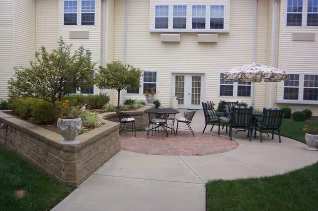 Outdoor patio area at River Pointe Health Campus featuring a paved circular seating area with metal tables and chairs, one table shaded by a floral umbrella. Raised garden beds with small trees and plants surround the patio, adjacent to a light yellow building with multiple windows and a double door.