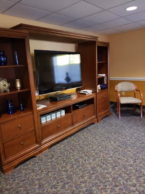 A cozy living room area with a large wooden entertainment center holding a flat-screen TV, decorative blue vases, and a set of blocks spelling 'FAITH'. To the right, there is a single upholstered chair with wooden arms. The room has beige walls and patterned carpet flooring.