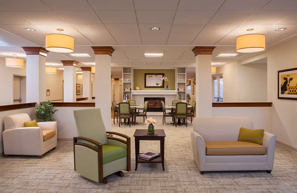 A spacious and well-lit living room area in a senior living facility featuring comfortable armchairs and a loveseat with green and beige cushions. In the background, there are dining tables with chairs arranged around a fireplace, flanked by built-in shelves with decorative items. The room has beige walls, carpeted flooring, and ceiling lights with drum shades.