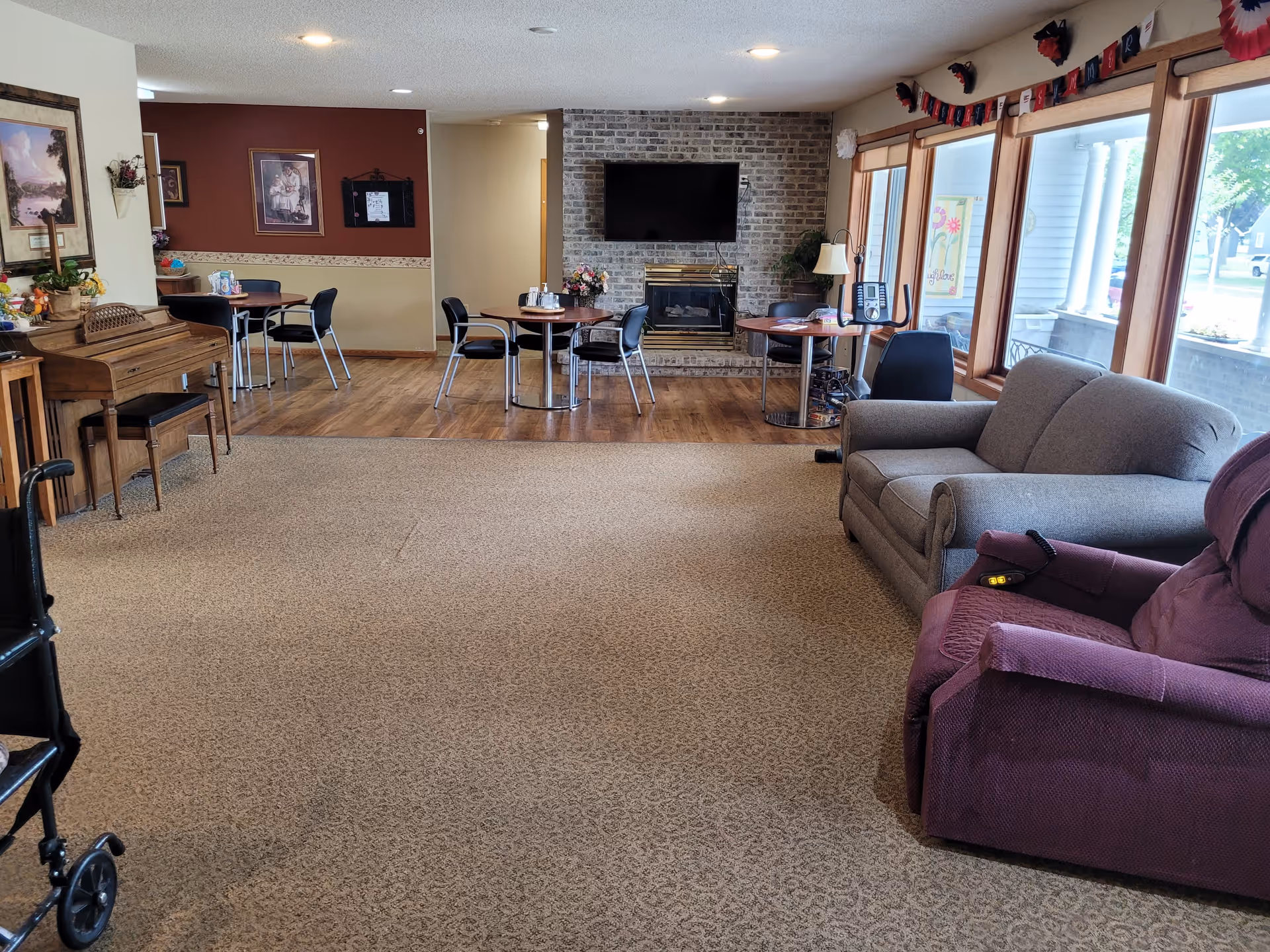 A senior living facility common area with a carpeted floor and a section of hardwood flooring. There are several round tables with chairs, a brick fireplace with a mounted TV above it, a gray couch, a purple recliner, and a wooden piano. Large windows let in natural light, and there is an exercise bike near the windows. The walls have framed pictures and decorations.