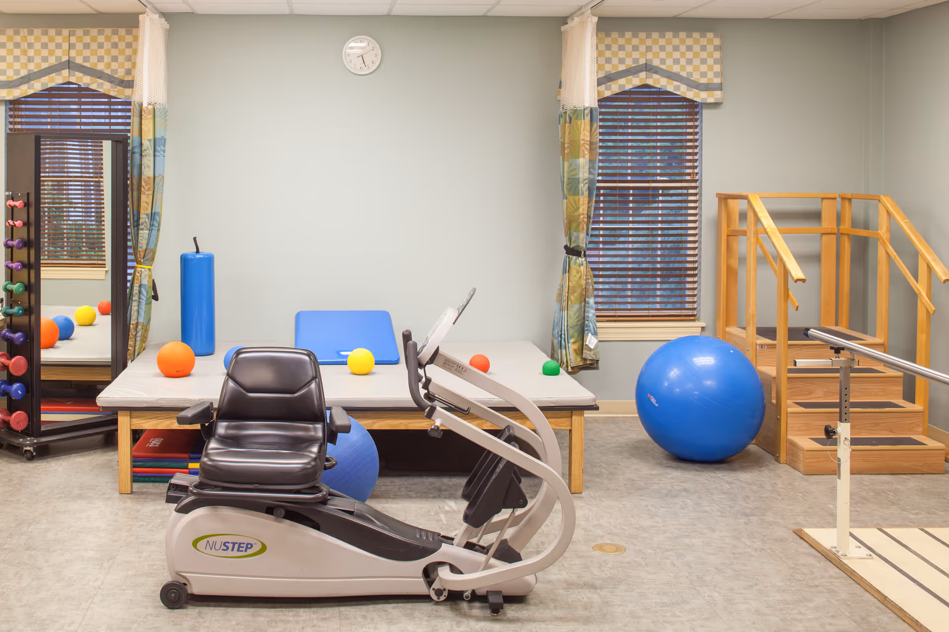 A physical therapy or exercise room with a recumbent exercise bike in the foreground, a large blue exercise ball, wooden stairs with handrails, a padded therapy table with colorful small exercise balls on it, a mirror, and windows with patterned curtains.