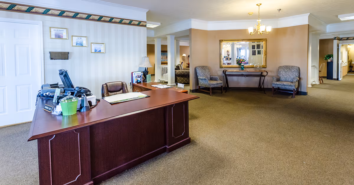 Reception desk and seating area in a senior living facility lobby with chairs, mirror, and a hallway.
