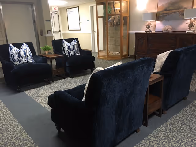 A cozy seating area in a senior living facility with four dark blue upholstered armchairs arranged around two wooden side tables. The chairs have decorative pillows, and the area is carpeted with a patterned rug. In the background, there is a wooden cabinet with a lamp and decorative items, a glass display case, and a hallway with exit signs.