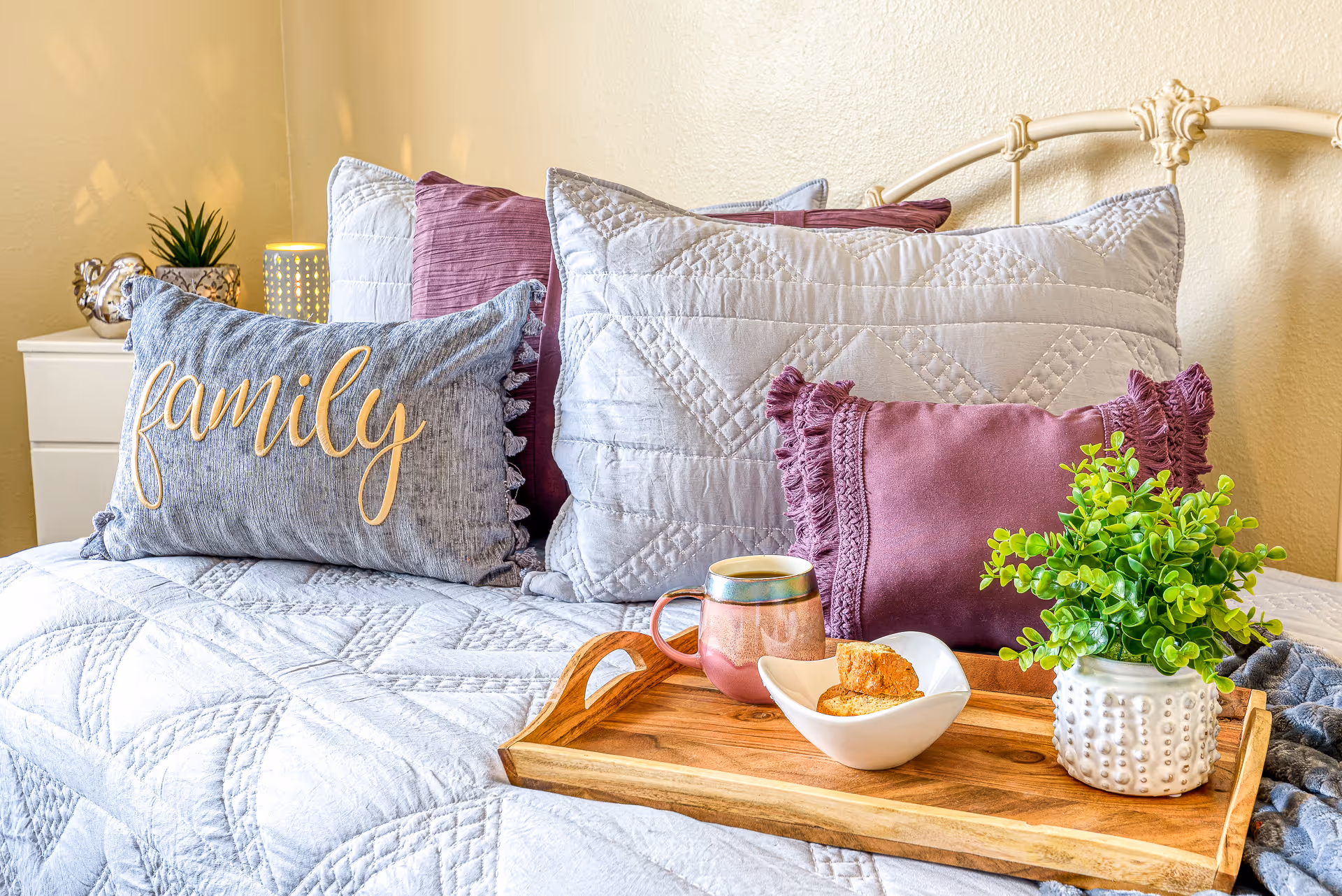 A neatly made bed with gray and purple decorative pillows (one embroidered with 'family') and a wooden tray holding a mug, a bowl of snacks, and a small potted plant.