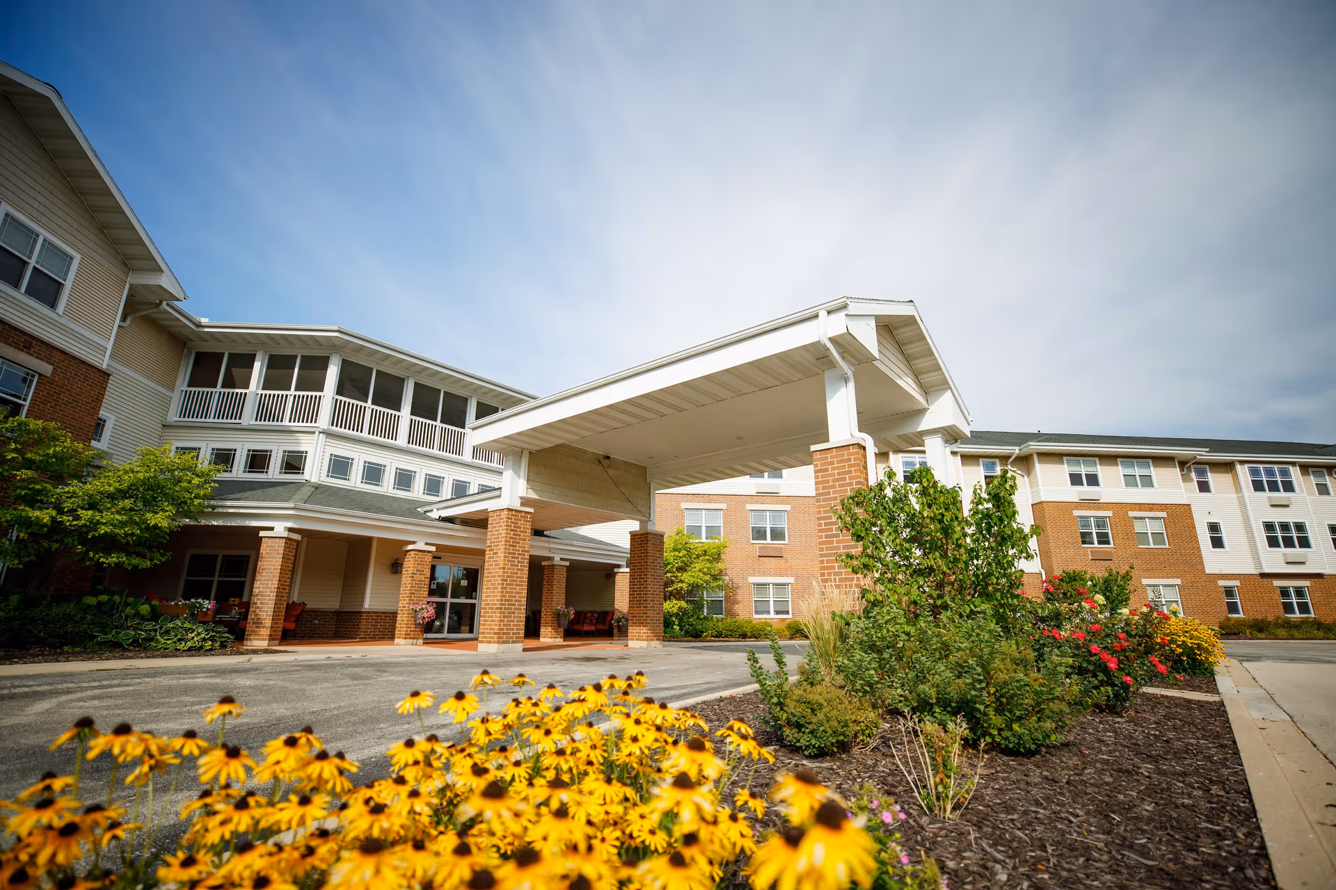 Exterior view of Hickory Park Senior Living building with a covered entrance, brick and beige siding, surrounded by landscaped flower beds with yellow and red flowers under a partly cloudy sky.