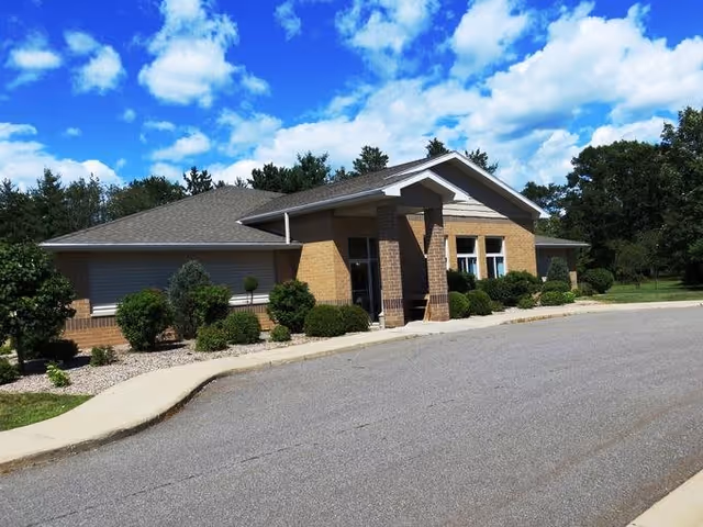 Exterior view of a single-story brick building with a gabled roof, surrounded by neatly trimmed bushes and trees under a bright blue sky with scattered clouds. A paved driveway and sidewalk lead up to the entrance.