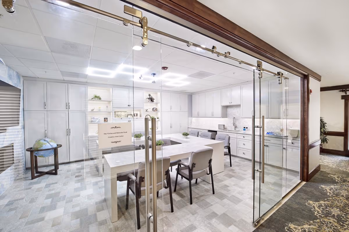 A bright and modern creative arts center room with white cabinetry, a large U-shaped table with chairs, and glass sliding doors. The room has a clean, organized look with decorative plants and shelves displaying various items. A globe on a stand is visible to the left outside the room.