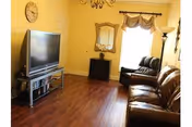 A living room with wooden flooring, a large flat-screen TV on a metal stand with shelves, a brown leather sofa, a matching leather recliner chair near a window with a beige valance, a floor lamp, a wall clock, a mirror above a small black cabinet, and yellow painted walls.