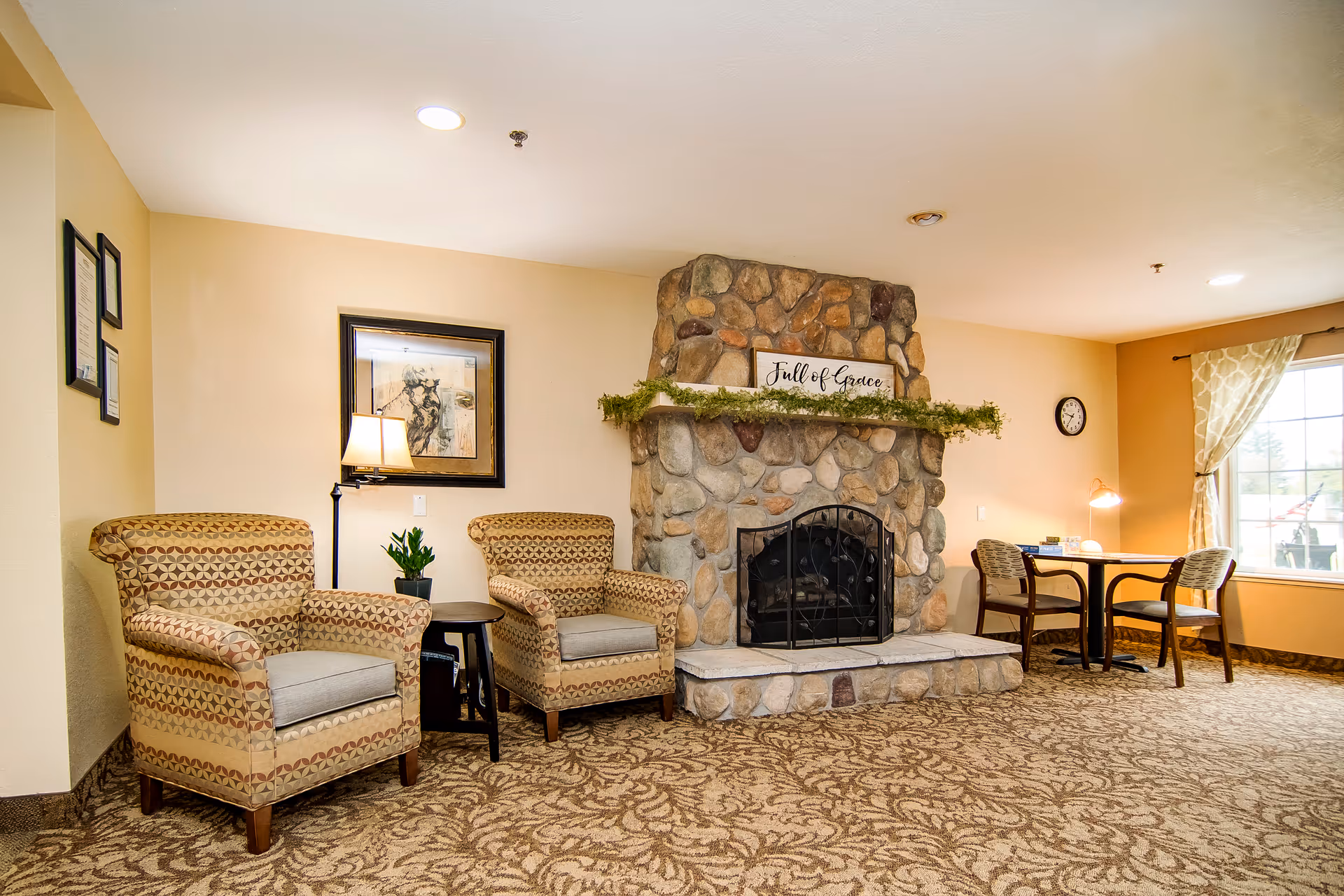 A cozy living room area featuring two patterned armchairs with a small round table and lamp between them, a stone fireplace with a decorative sign reading 'Full of Grace' and greenery on the mantel, and a small table with two chairs near a window with patterned curtains. The room has beige walls and patterned carpet.