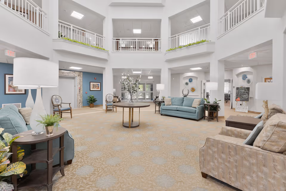 Spacious and well-lit living room area in a senior living facility with beige patterned carpet, light blue and beige sofas, wooden side tables with lamps and plants, a round table with a flower arrangement in the center, and a second-floor balcony with white railings overlooking the room.