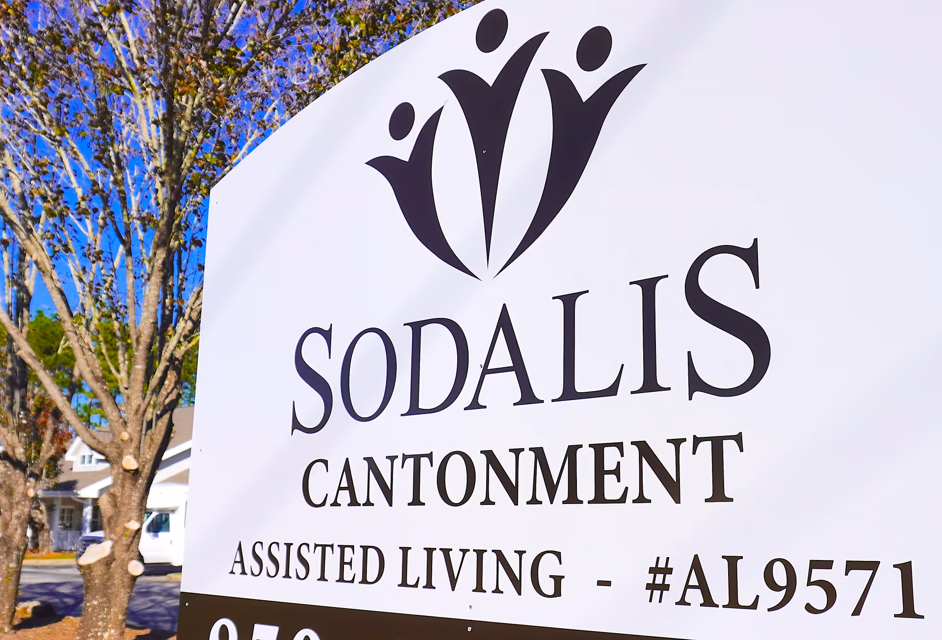 Close-up of a white sign for Sodalis Cantonment Assisted Living with a logo of three stylized human figures above the text. Trees and part of a building are visible in the background.