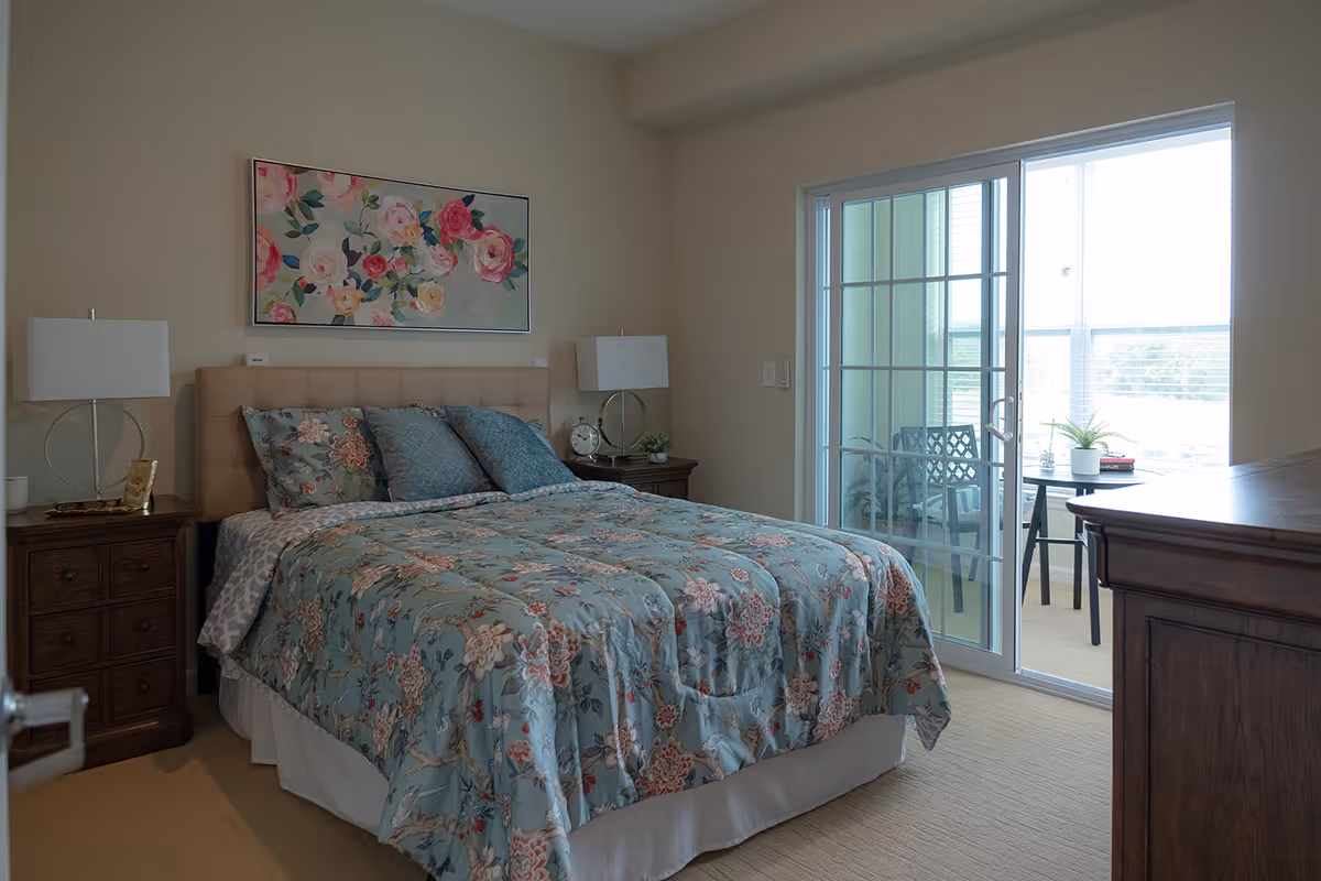 A bedroom with a bed covered in a floral patterned comforter and pillows. There are two bedside tables with lamps on each side of the bed. Above the bed hangs a painting of colorful flowers. To the right, there is a sliding glass door leading to a small balcony with a table, chairs, and a potted plant.