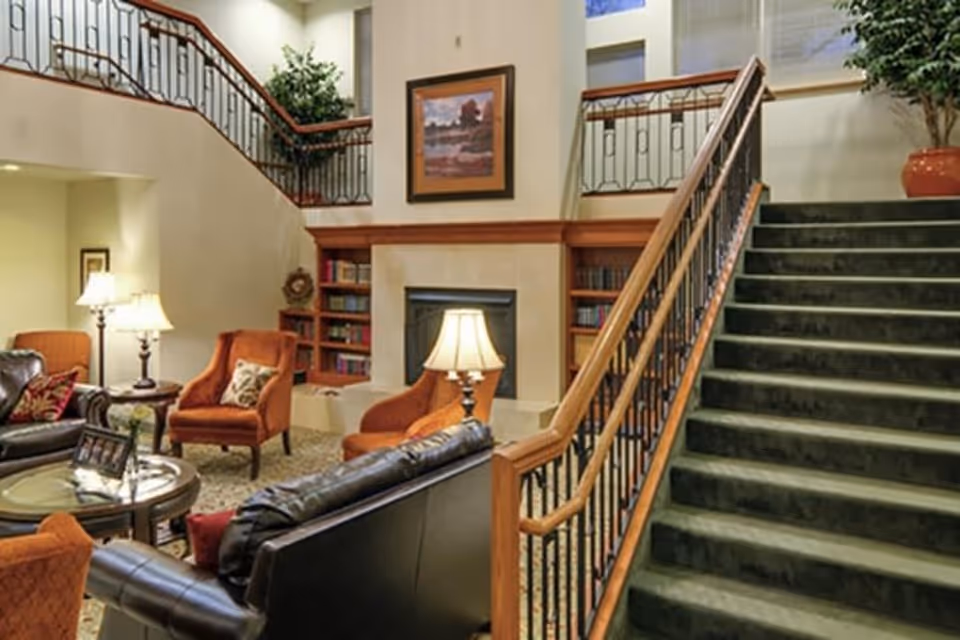 A cozy living room area in Cherry Park Plaza featuring a staircase with a wooden handrail and green carpet. The room includes a fireplace with built-in bookshelves on either side, several armchairs and a leather sofa arranged around a glass coffee table, and a table lamp providing warm lighting. There is a framed painting above the fireplace and a large potted plant near the staircase.
