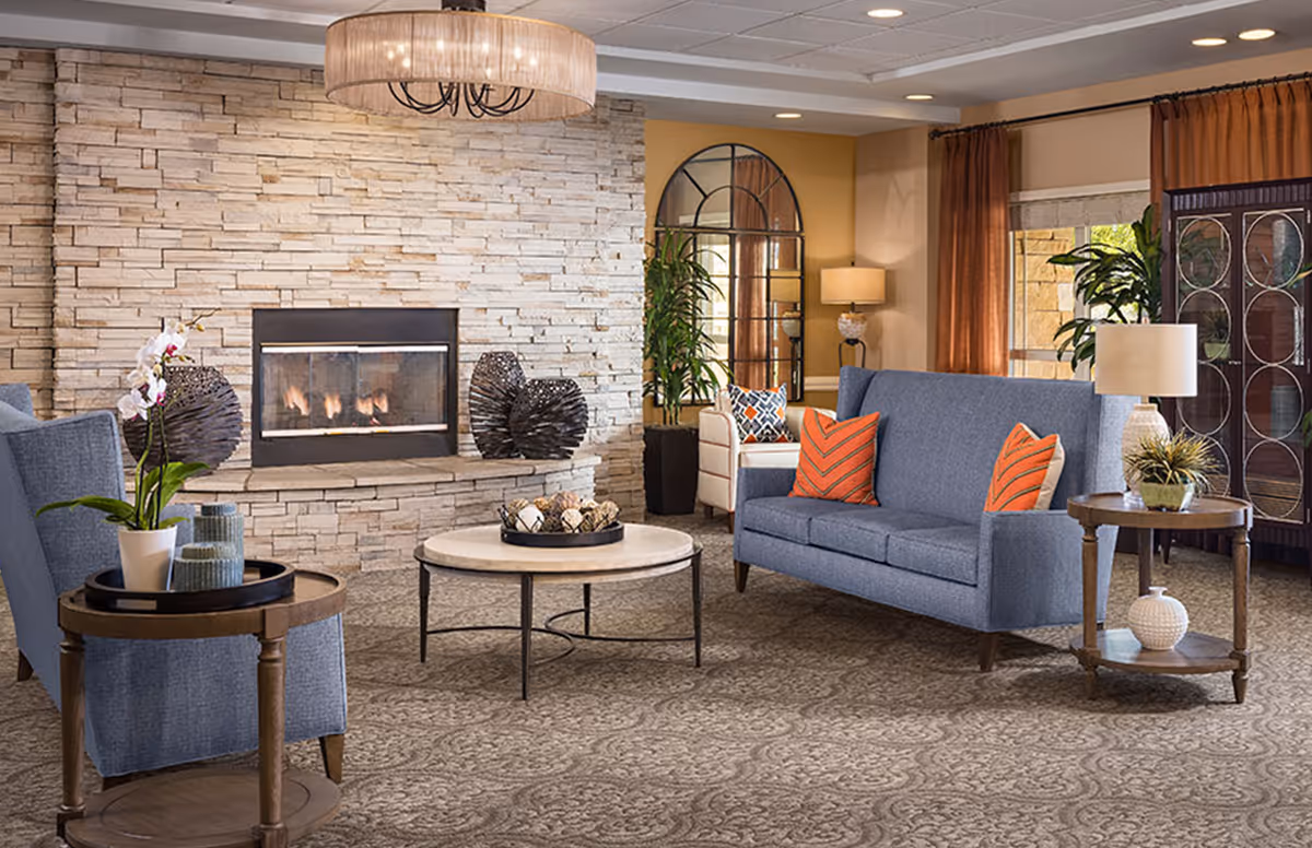 A cozy senior living facility lounge area with a stone fireplace, a blue sofa with orange pillows, two blue armchairs, round wooden side tables with plants and decorative items, a round coffee table with decorative balls, a large mirror, and warm lighting.