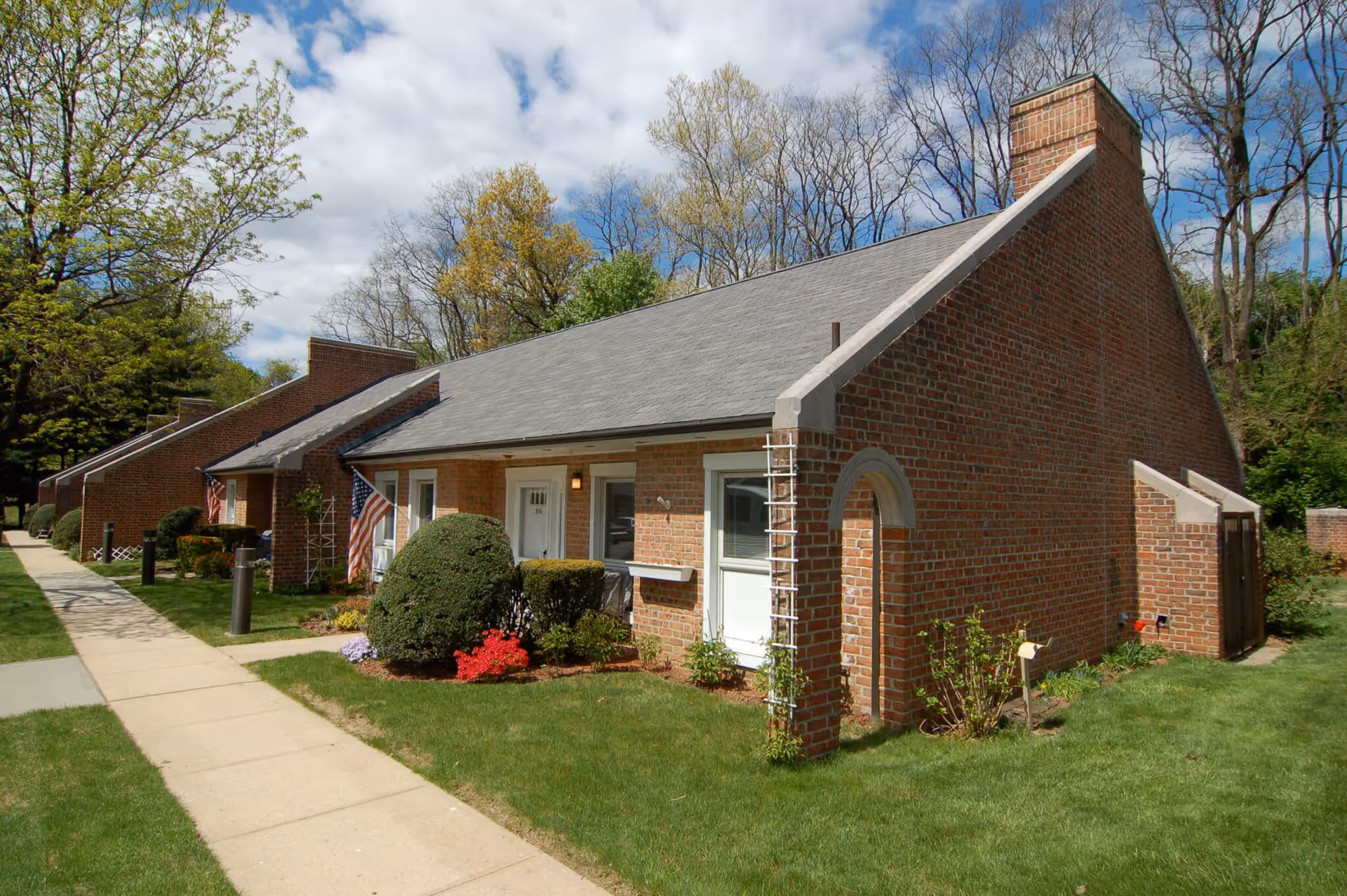 A row of single-story brick residential buildings with sloped roofs, small front yards with bushes and flowers, and American flags near the entrances. A concrete sidewalk runs alongside the buildings, and trees with early spring foliage are visible in the background under a partly cloudy sky.