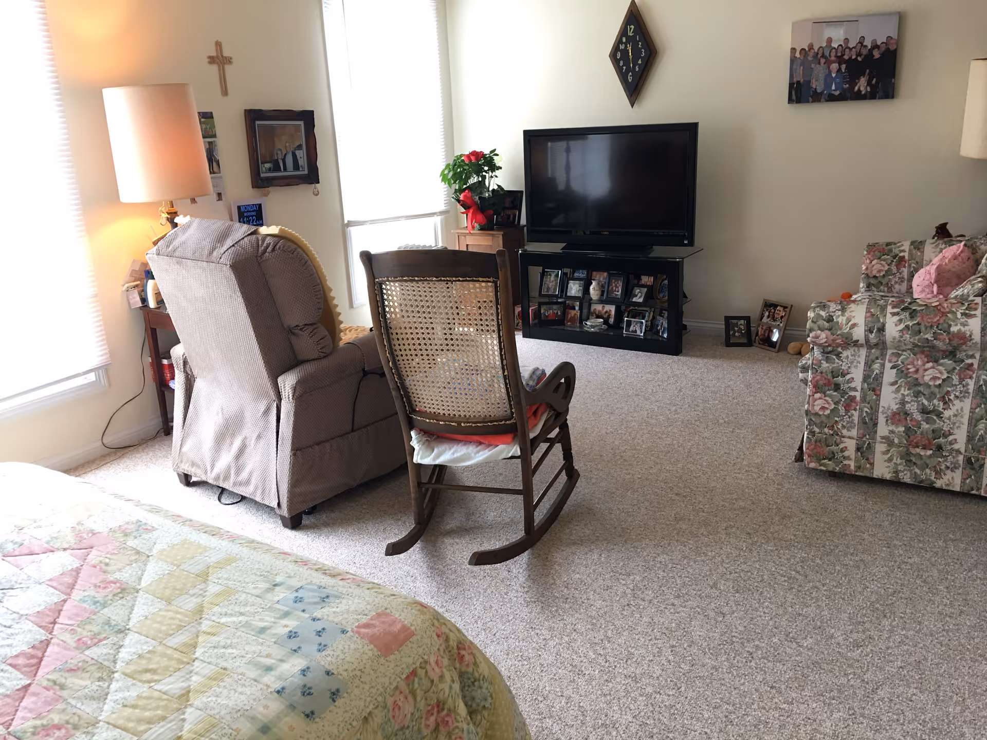 A cozy living room area with a beige recliner chair, a wooden rocking chair with a cushion, and a floral patterned armchair. There is a TV on a black stand with several framed photos underneath. A lamp with a beige shade is on a side table next to the recliner. The room has light-colored carpet and walls, with a cross and a clock hanging on the wall. A quilted bedspread is partially visible in the foreground.