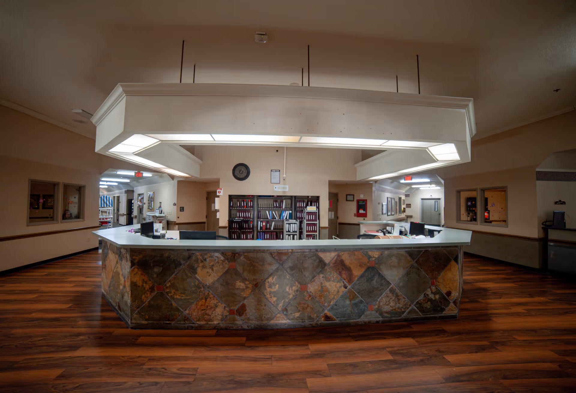 A wide reception desk area in a healthcare facility with a tiled front and overhead fluorescent lighting. Behind the desk are shelves filled with binders and office supplies. The floor is wood-patterned, and the walls are beige with some framed items and windows into adjacent rooms.