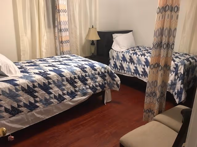Small bedroom with two twin beds covered in blue patterned quilts, a nightstand with a lamp, curtains and a small bench.