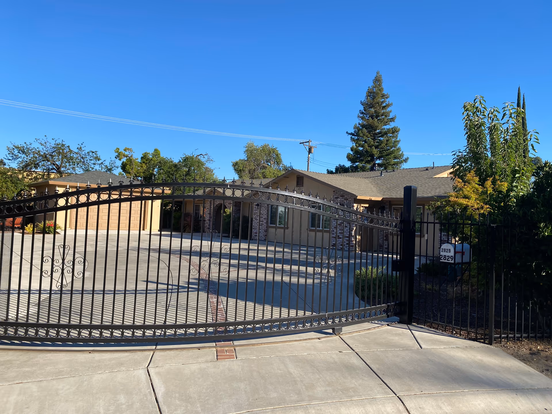 View of a single-story residential building behind a black wrought iron gate with a concrete driveway and some trees and bushes around the property under a clear blue sky.
