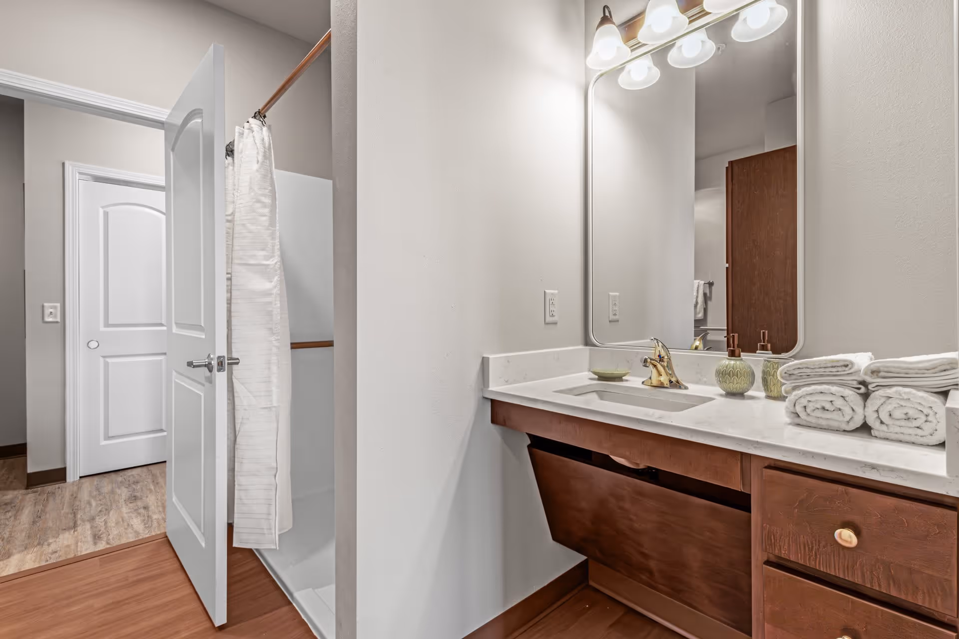 A bathroom with a white door partially open revealing a shower with a white curtain. The bathroom has a countertop with a sink, a large mirror, and several neatly rolled white towels. The cabinetry is wooden with a dark finish, and the floor is a wood-like material.