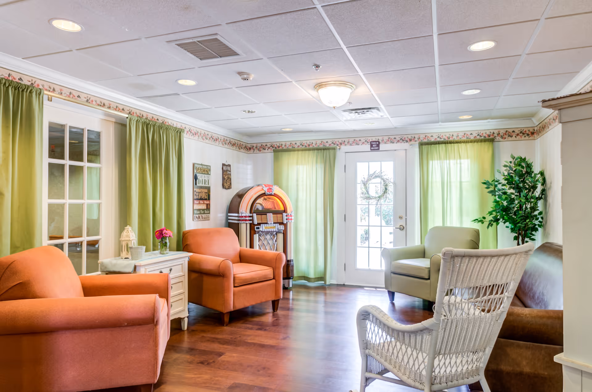 A cozy living room area with two orange armchairs, a white wicker chair, a green armchair, and a brown leather sofa. The room features green curtains, a jukebox in the corner, a small white side table with a flower vase, and a potted plant near a glass door with a wreath. The walls have floral wallpaper border near the ceiling and wooden flooring.
