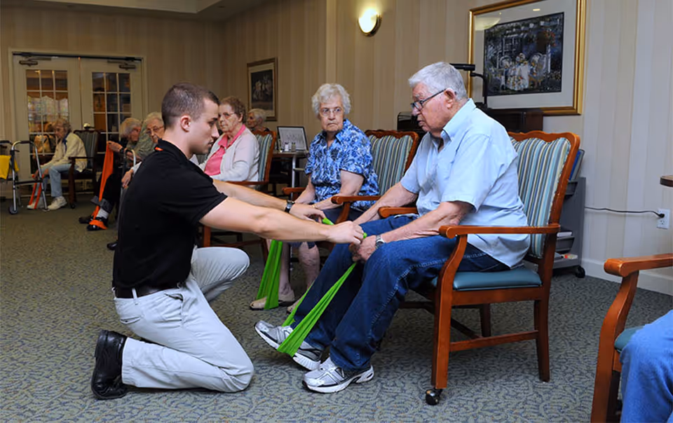 A young male caregiver kneels on the floor assisting an elderly man seated in a chair with leg exercises using a green resistance band. Other elderly individuals are seated in the background in a room with beige walls and carpeted floor.