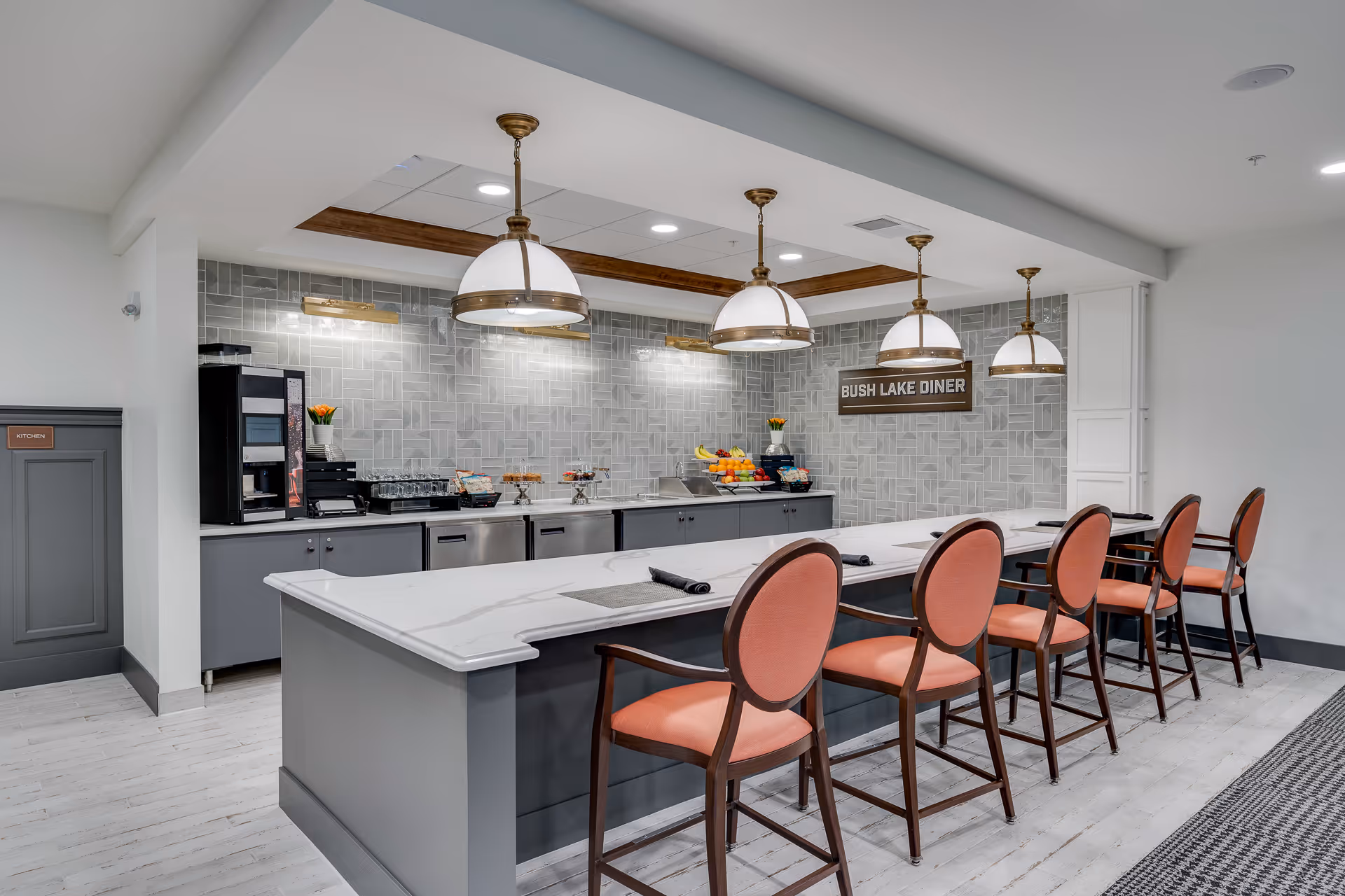Interior view of a modern dining area named Bush Lake Diner with a long white countertop island and six high-back chairs with coral upholstery. The back wall features a gray tiled backsplash, a coffee machine, glassware, fruit bowls, and pendant lights hanging from the ceiling.
