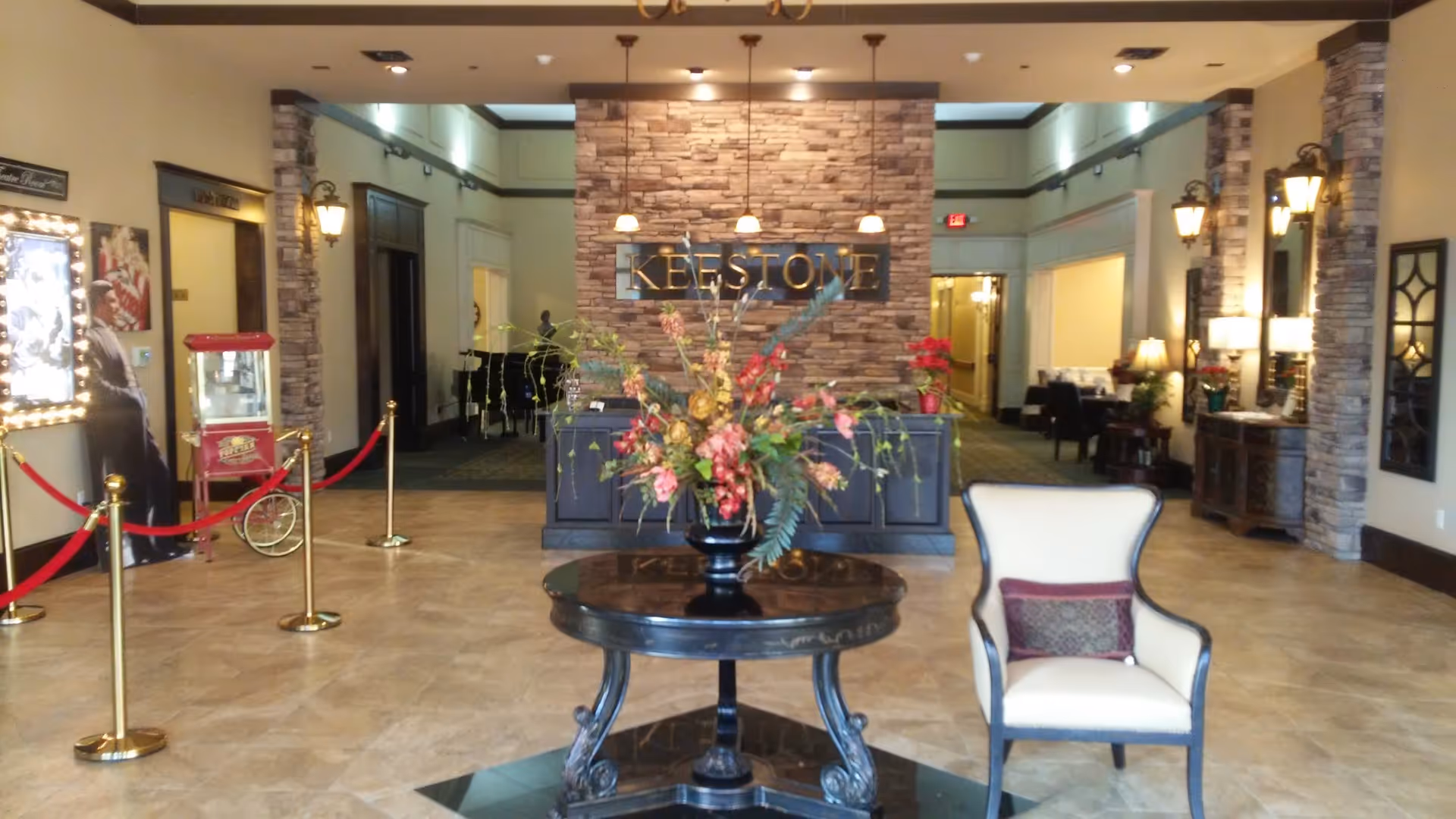 Lobby area of Keestone at Hewitt House featuring a round table with a large floral arrangement in the center, a single upholstered armchair with a decorative pillow to the right, and a stone accent wall behind the reception desk with the word 'KEESTONE' displayed. The space has tiled flooring, warm lighting fixtures, and decorative elements including a popcorn machine and framed pictures on the left side.