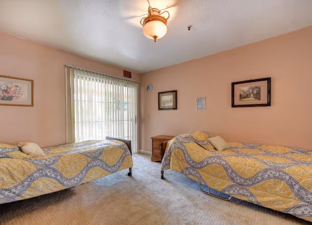 A bedroom with two twin beds covered in yellow and gray patterned bedspreads. There is a wooden nightstand between the beds, and three framed pictures hang on the peach-colored walls. A sliding glass door with vertical blinds leads outside, and a ceiling light fixture is visible.