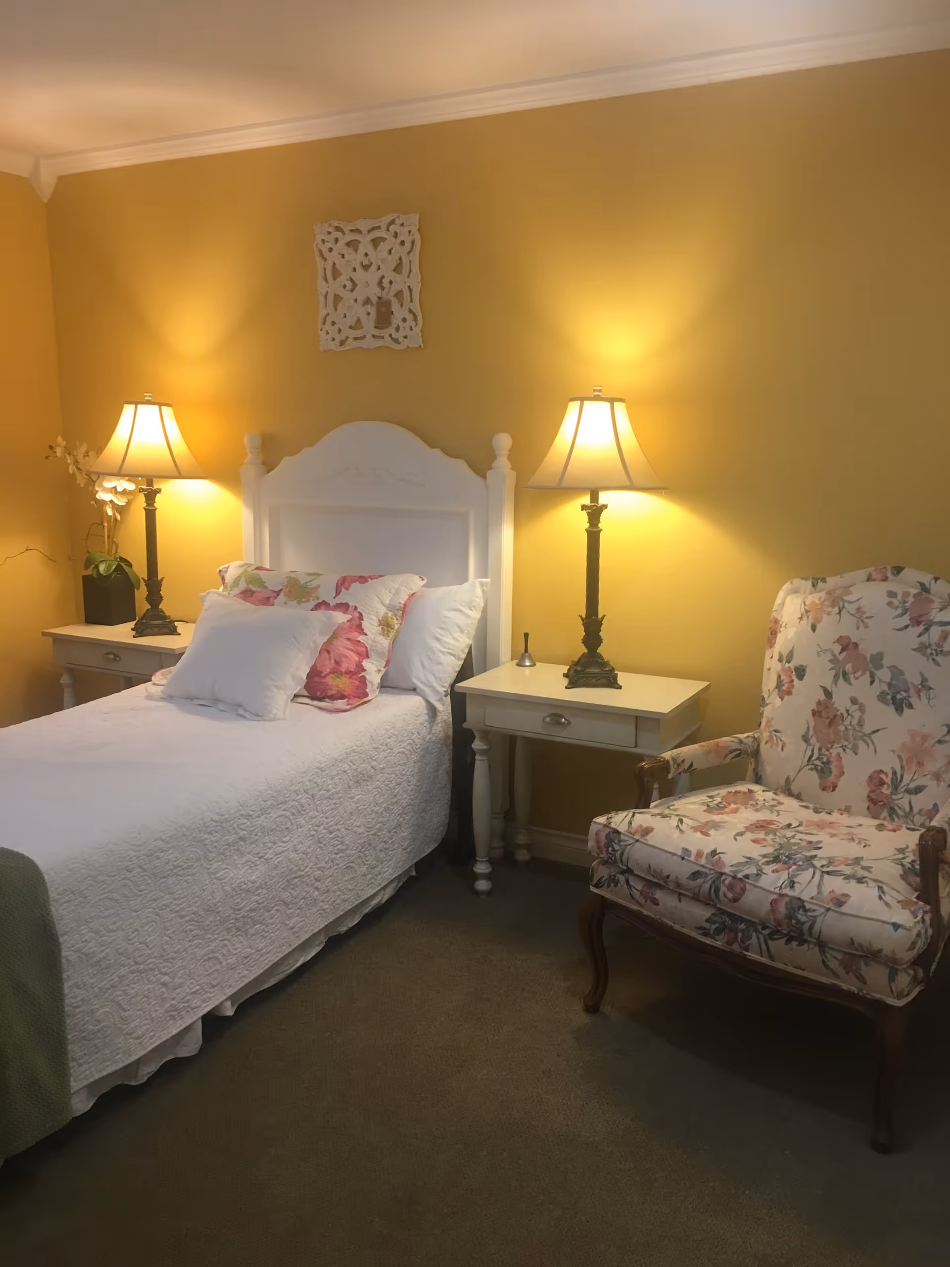 A cozy bedroom with a single white bed adorned with floral pillows and a white quilt. On either side of the bed are white nightstands, each with a lamp emitting warm light. To the right of the bed is a floral upholstered armchair. The walls are painted yellow and there is a decorative white wall hanging above the bed.