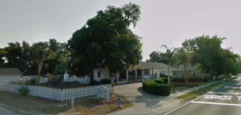 Single-story senior living facility with white picket fence, trees, and a curved driveway.